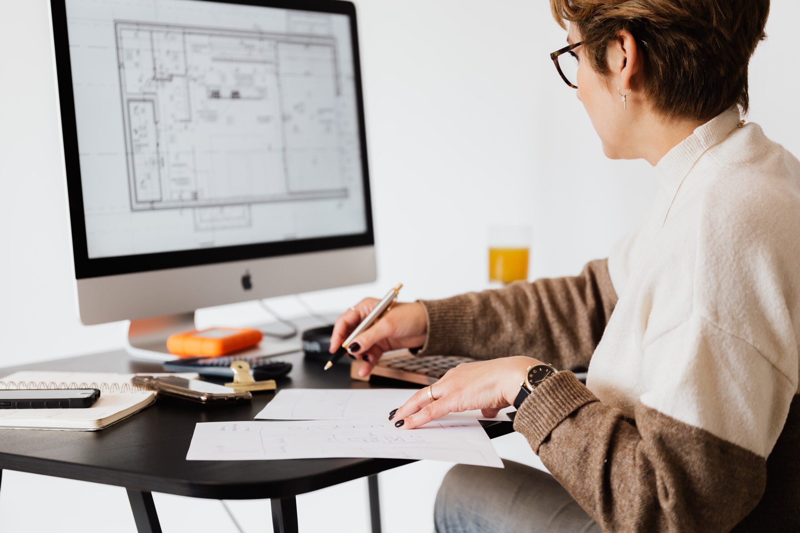Profissional de Design de Interiores olhando planta de projeto no computador e com um papel e uma caneta na mão